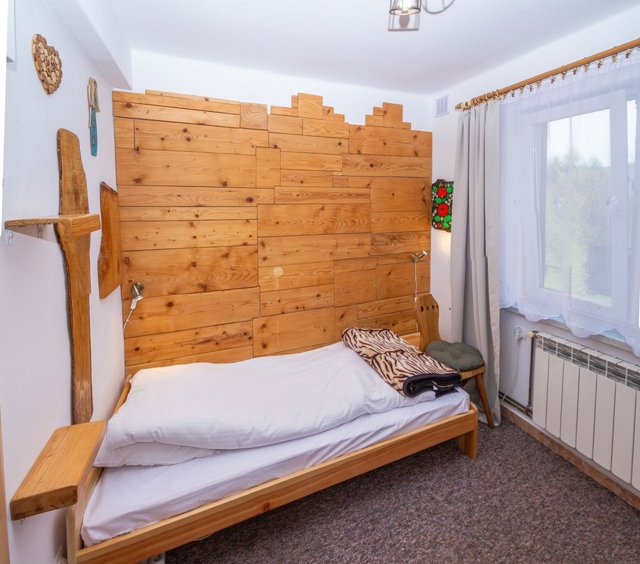 Pokój z drewnianą ścianą za łóżkiem, oknem z firaną i widokiem na zieleń