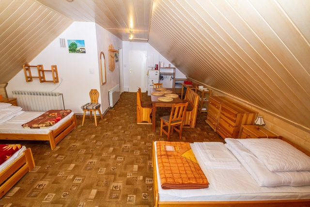 Apartament na poddaszu z drewnianym sufitem: cztery pojedyncze łóżka, dwa złączone razem, stół z krzesłami i aneks kuchenny w głębi